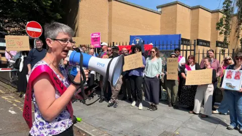Maggie Chapman, a woman with short grey hair and glasses addresses a protest through a loudhailer. A crowd of people holding banners are in the background.