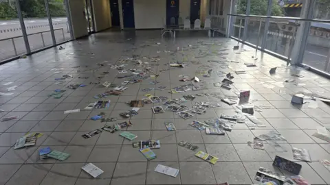 Ipswich Police An interior photo of a bus terminal. Leaflets are scattered on the, there are tyre skid marks on the floor. The building has glass windows either side and seating at the back.