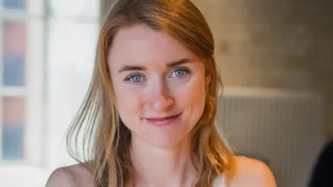 Woman with mouse coloured hair and blue eyes smiling at the camera. She is standing inside a room with a window in the background.