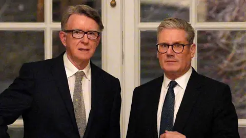 Peter Mandelson stands with Sir Keir Starmer in front of a set of double doors with white panes. Both men wear black suit jackets, white shirts and dark ties.