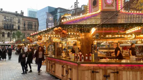 BBC/Phil Cunliffe A wooden Manchester Christmas market hut covered in fairy lights with people inside cooking hotdogs. There are ketchup and mustard containers on the counter and members of the public are walking past.