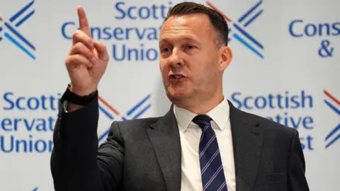 Russell Findlay wearing a dark suit over a white shirt and blue and red striped tie while pointing in the air and standing in front of a white background with the words Scottish Conservative and Unionist on it.