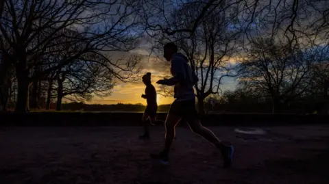 PA Media Silhoutted joggers run through a park. The sun is low in the sky, and there are trees surrounding the runners