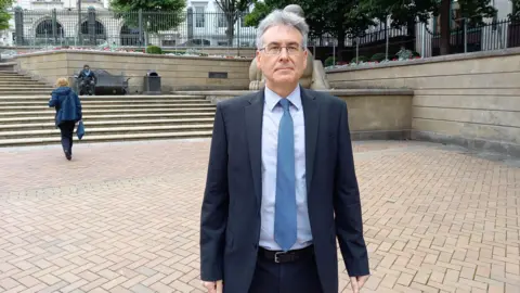 LDRS Simon Foster standing in the middle of a town square with steps behind him. He is wearing glasses, a suit and a blue tie. He has short, grey hair and his arms are at his sides. 