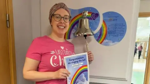 Laura Rush Laura is wearing a pink t-shirt and a patterned head turban, and is wearing glasses. She is smiling and has her left hand on the rope of a bell attached to a wall. In her right hand she has a certificate with a rainbow on it and it says "congratulations on ringing the bell".