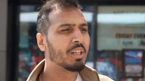 Satwinder Sony is a young asian man, with dark hair and a dark beard, he is wearing a brown coat with a high collar. 