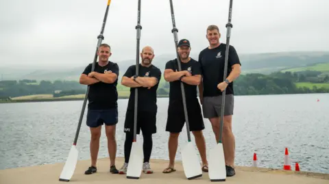BBC Four men in T-shirts and shorts stand on the edge of a lake carrying large oars.