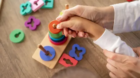 Getty Images A close-up image of an adult helping a young child place a colourful round piece onto a wooden peg toy. The toy consists of multiple pegs and various brightly coloured, differently shaped pieces scattered around on a wooden surface.
