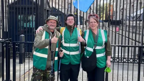 Rob, Lisa and son Drew outside gates in Downing Street. They are wearing dark green jackets and Rob Collins on the left is putting his thumb up. 