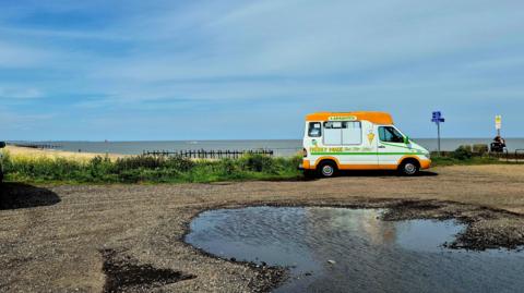 An ice cream van on a damp coast under a sky with thin cloud