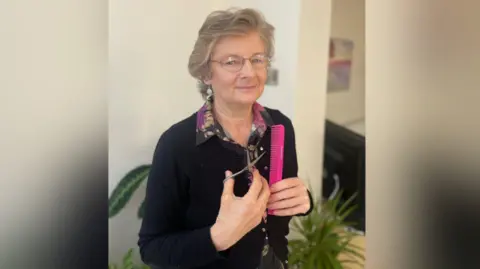 Jenny Wells Petition initiator Jenny Wells. She is smiling for the camera and holding a magenta comb and a pair of scissors. There are plants in pots behind her.