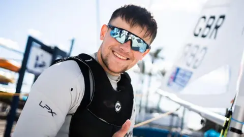 Arthur is smiling at the camera. He has light blue reflective sunglasses on and wears a black waterproof vest with a light grey long sleeved wetsuit. The background is slightly blurred but the sail of a Team GBR yacht can be seen.