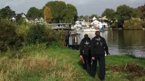Two uniform-clad enforcement officers walk along the riverbank towards boats.
