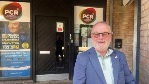 Wayne Fitzgerald wearing a denim coloured blue suit and a polo shirt in pale green is smiling outside the officers of PCRFM radio station. The windows are covered with posters advertising the ratio station output.

