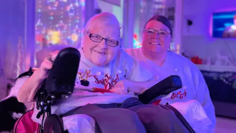 Sandra Lee Myles and her mum Liz smile at the camera in the living room. They are wearing matching Christmas jumpers and there are different Christmas decorations and lights in the background.