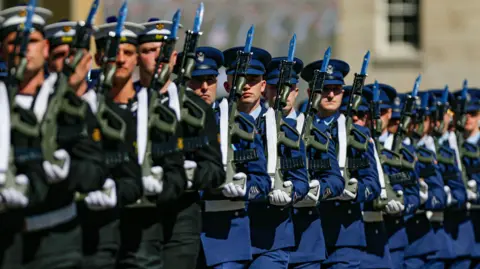 Defence Forces members on parade in green and blue uniforms, holding weapons in air