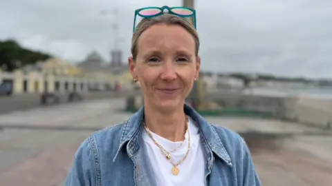 Lorna Gleave has fair hair tied back, with sunglasses resting on her head. She's wearing a white t-shirt and a denim jacket.