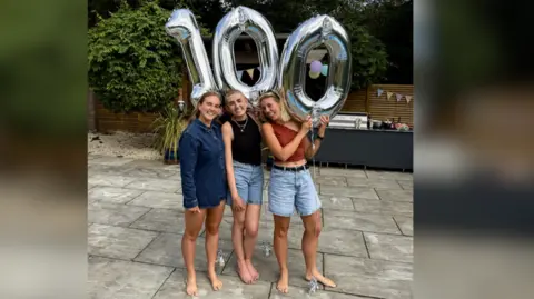 Alice Boston The image shows three women standing barefoot outside holding a three silver balloons to make the number 100.