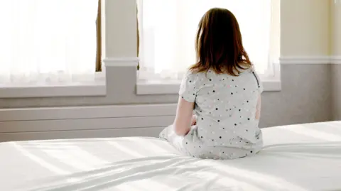 Young girl in nightdress sits on a bed with a white sheet with her back to the camera and looks out the window.