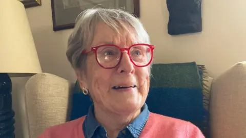 A woman sits in an armchair looking to the right. She has red glasses, grey hair and a salmon-coloured cardigan. She is smiling.