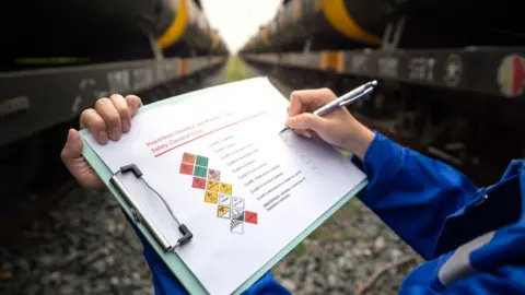 A person holding a clipboard with a pen, writing on it. The paper on the clipboard has various hazardous symbols on with a bullet point check list next to it. Freight trains in a yard can be seen behind them. 