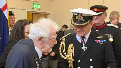 GUY CAMPBELL/BBC Jim Grant is wearing a suit and glasses and talking to Robert Bellfield who is wearing a formal military jacket with golden braid attached and a Royal Navy officer cap