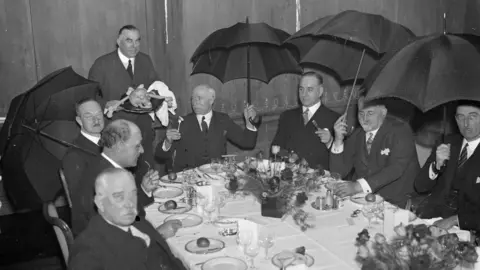 Alamy Archive photo from 1934 showing men sitting at an elaborately-decorated dining table while holding open umbrellas above their heads