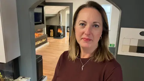 A woman with long blonde hair and wearing a silver necklace and burgundy top, stands and looks at the camera. She is inside a shop which sells stoves and fire places.