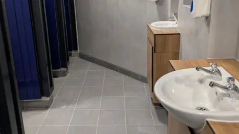 A clean, modern public restroom with a tiled grey floor and smooth light‑grey walls. Along the right side, there are two white sinks mounted on wooden cabinets, each fitted with chrome taps. A soap dispenser is mounted on the wall above the sinks. On the left side of the image, several cubicle doors in a dark blue colour are visible.

