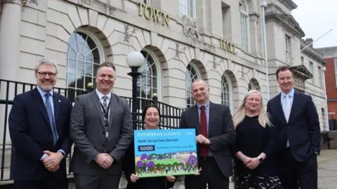 Four men and two women are standing in a line smiling at the camera. A woman and man n the middle are holding up a small posted which says "ROTHERHAM COUNCIL Adult social care services GOOD" They are standing in front of Rotherham Town Hall which is an off-white stone building with large arched windows.