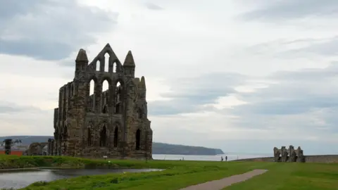 BBC A dramatic landscape featuring the ruins of a large stone abbey situated on a grassy headland overlooking the sea.
