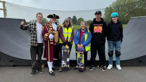 Royal Wootton Bassett Town Council Six people are pictured standing in front of a half-pipe. One man is wearing mayoral garb and is holding a hand-bell white two women are holding skateboards in front of them. There is a park behind the half-pipe and it appears to be a grey day.