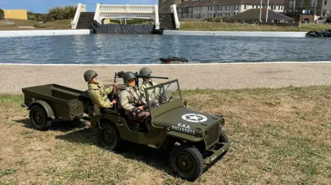 Redcar Model Boat Club A model khaki-green USA military vehicle with three model soldiers travelling in it. Park of the refilled lake is in the background with a model boat sailing in it. An overhead bridge rises above the centre of the lake, splitting it in two. There are houses further away and the one-storey building of nearby cafe Boathouse.