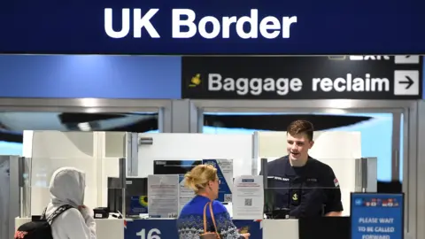 PA Media Two people queue at the UK border at Heathrow Airport in London. One woman is wearing a blue patterned jumped and has a handbag on her shoulder. The border agent, a man with dark hair, is smiling. A third person, dressed in a grey hoodie has their face hidden.