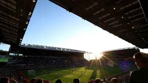 Getty Images A view of the inside of Villa Park when a football game is on. Thousands of people are sitting in four stands around a green football pitch with players on it. The sun is shining in the far corner.