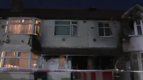 A view of a terraced-appearing house in Berkley Avenue in Hounslow with burnt windows and some police tape at the front