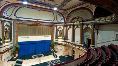 Ian Grundy The ornate interior of Tottenham Palace Theatre, showing rows of red seats facing a stage framed by decorative columns, sculptures and gilded detailing, beneath an elaborately decorated ceiling and curved balcony.