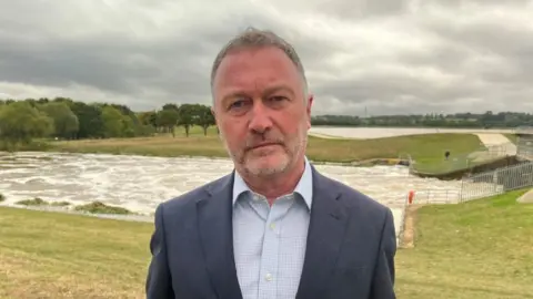 Steve Reed with short grey hair and beard, wearing a grey jacket and striped shirt stands in front of a flooded field