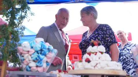 PA Media King Charles talks to a woman in a patterned dress in front of a tower of meringues