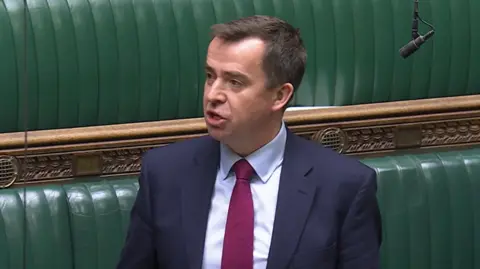 Parliament TV Calum Miller speaks against the backdrop of the green leather benches of the House of Commons. He has dark hair with a parting and wears a dark blue suit, a white shirt and a mauve tie.