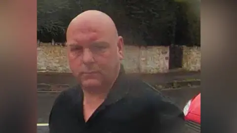 Man with black top on a street with a wall and bush behind him and a red car.