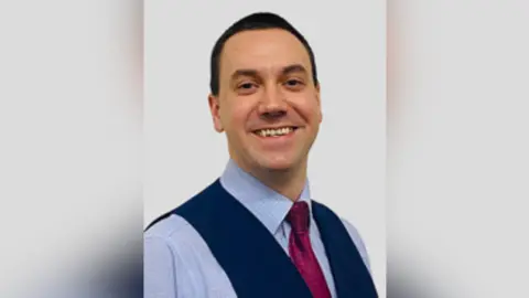 Alastair Redman looking directly at the camera. He has dark hair and is smiling. He is standing against a neutral background. He is wearing a blue shirt with a red tie and a dark blue waistcoat over the top.