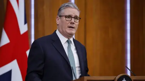 Getty Images Keir Starmer is standing in front of a microphone. There is a large Union Jack flag behind him. He is wearing dark framed glasses and has greying hair. He is wearing a navy suit, white shirt and green patterned tie.