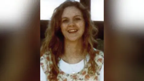 An Garda Síochána A woman with long, curly brown hair, smiling, she is wearing a white top with a floral pattern