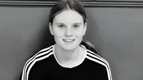 Family Brochure Black and white photograph of Holly. She is sitting with her back against a wall and smiles at the camera.