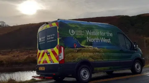 A United Utilities van parked on a road in the countryside with a river the left and hills in the background. The sun is peering out through a cloud.