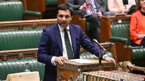 Saqib Bhatti is wearing a navy blue suit jacket with a white shirt and dark green tie. He is standing at a podium inside the House of Commons, with dark green long benches in the background. There is a microphone in front of him. 