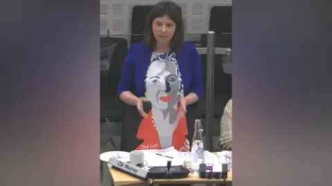 Suffolk County Council/YouTube Sam Murray is standing up in the council chamber at her desk. She is wearing a red, blue and grey dress with a picture of Margaret Thatcher's face down the front of the whole dress