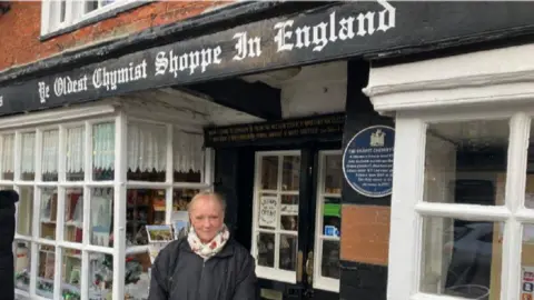 Carol Barden wearing a scarf and black coat stood outside Ye Oldest Chymist Shoppe in England