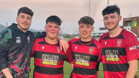 Family Photo There are four rugby players in this photograph, all of whom are young men. They're all wearing rugby shirts. The boys all have dark hair and they've got their arms around each other. 
